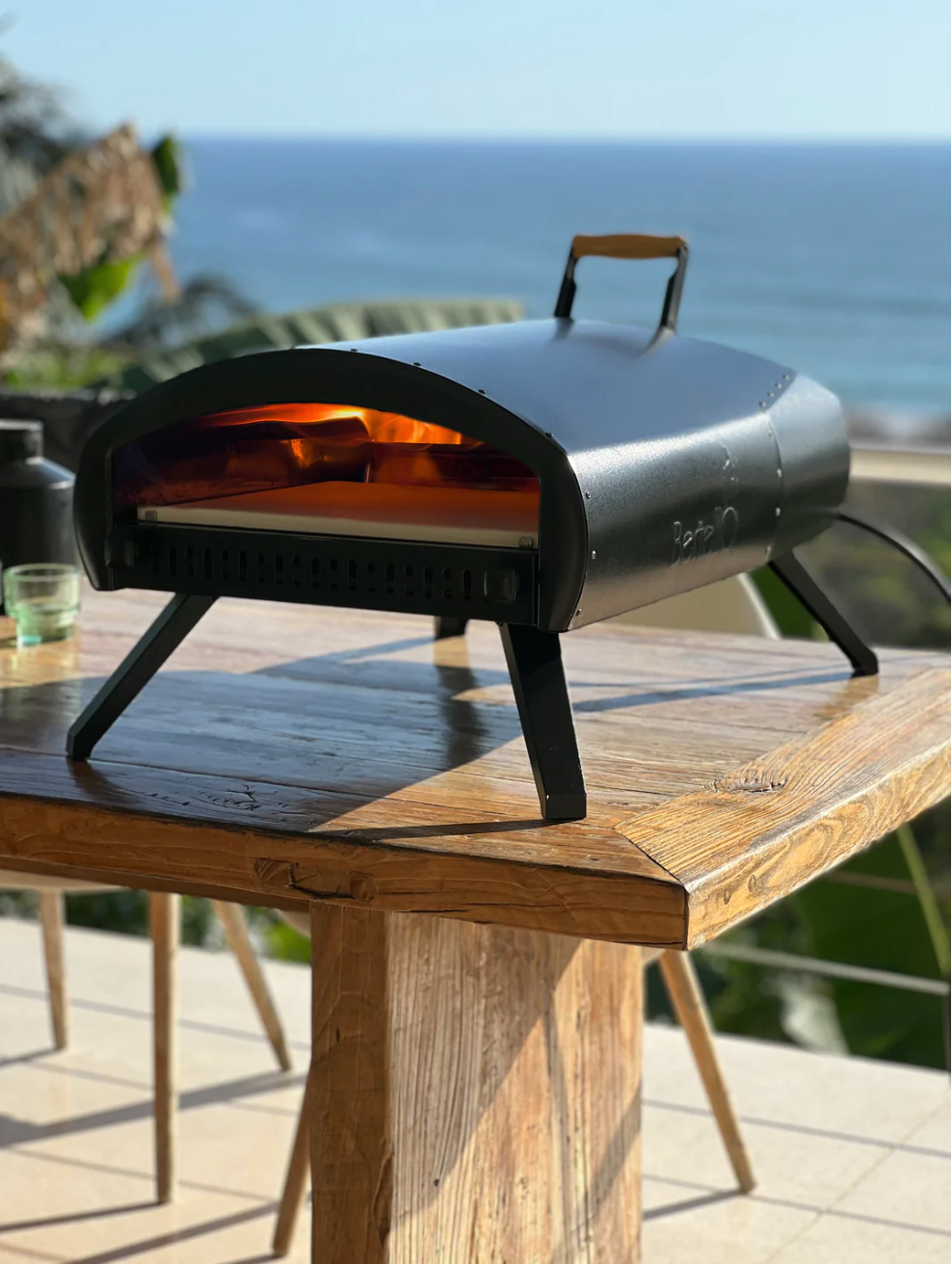 Bertello Grande 16-inch Non-Rotating outdoor pizza oven on legs on a wooden table, shown with its fire lit.